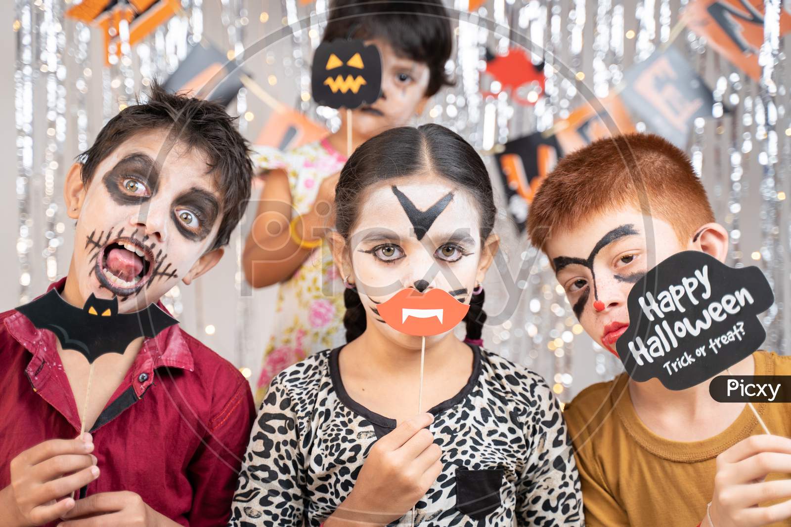 Group Of Kids In Halloween Costumes Making Gesticulating Scary Or Spooky Faces By Holding Booth Sprops On Decorated Background By Looking Into Camera.