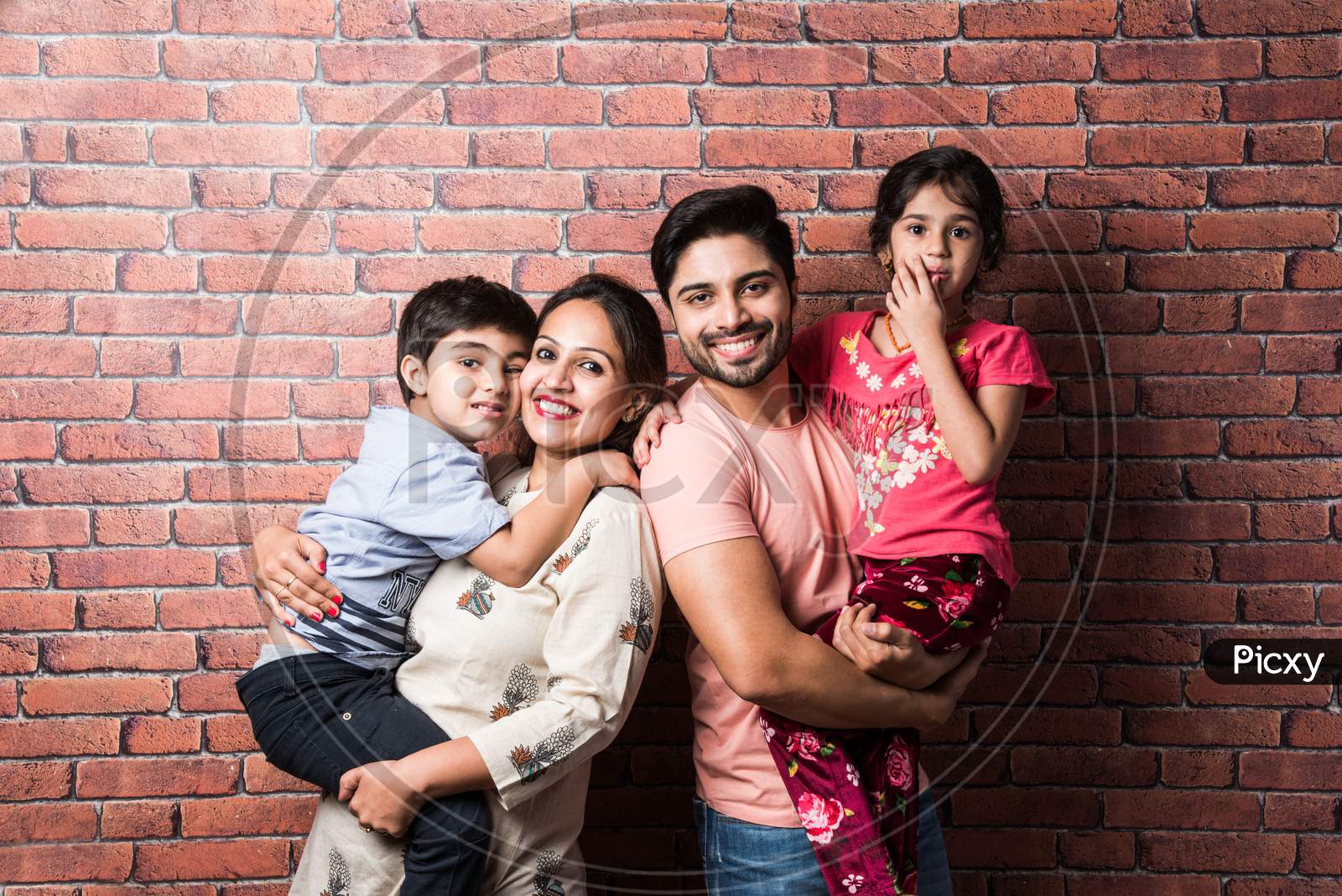 Beautiful Indian Young Family Of Four Hugging While Standing Against Red Brick Wall