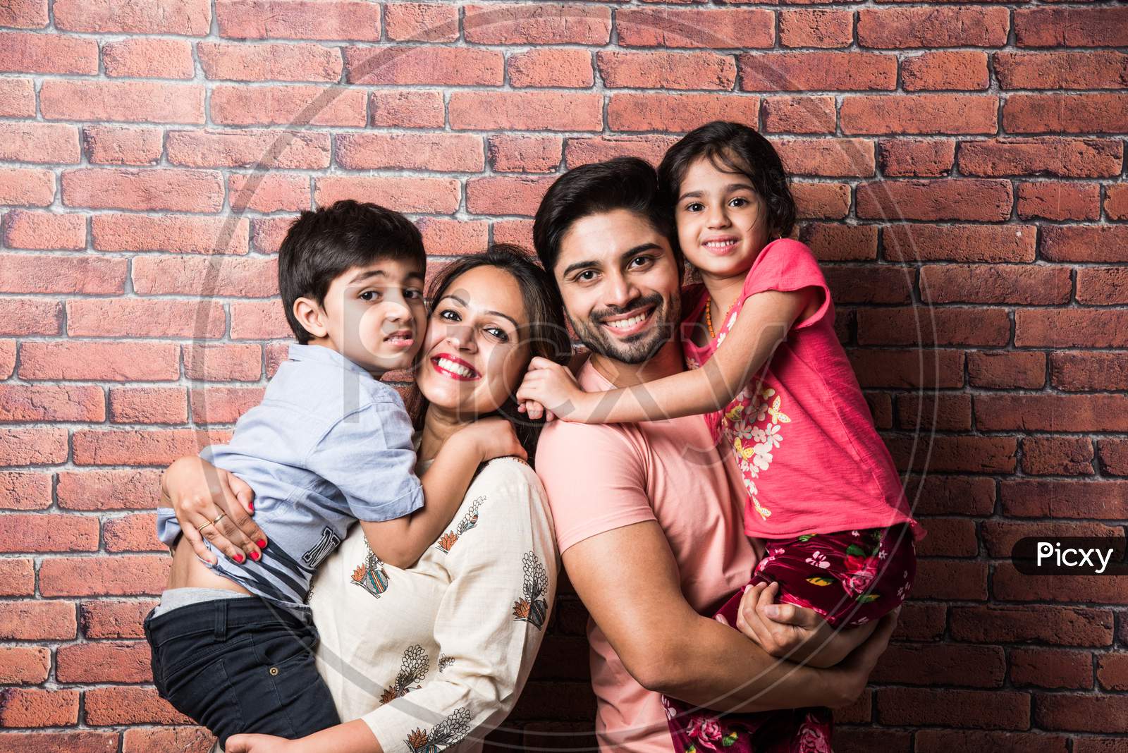 Beautiful Indian Young Family Of Four Hugging While Standing Against Red Brick Wall