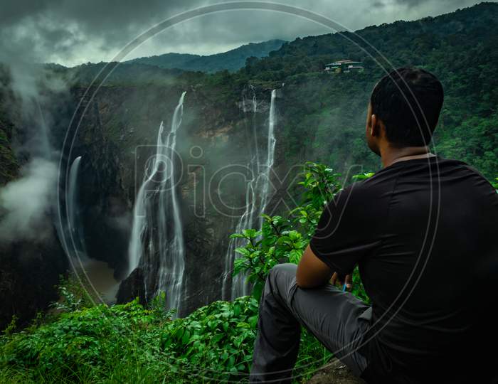 Man Sitting At Rock Watching The Beautiful Waterfall Stream At Evening