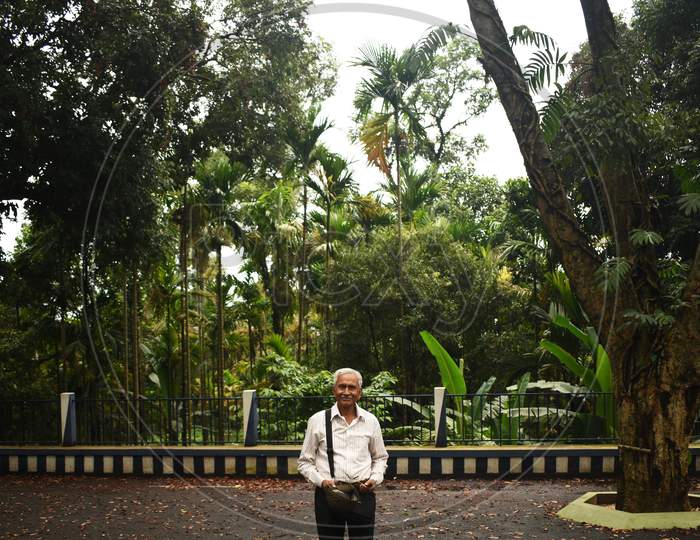 An Old Man, Wearing Spectacles And Shirt, Standing Alone In Mawlynnong Village, East Khasi Hills, Meghalaya, India. The Elder Person With Cap In Hand, Side Bag On Shoulder, Looking Towards The Camera.