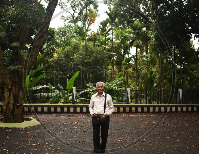 An Old Man, Wearing Spectacles And Shirt, Standing Alone In Mawlynnong Village, East Khasi Hills, Meghalaya, India. The Elder Person With Cap In Hand, Side Bag On Shoulder, Looking Towards The Camera.