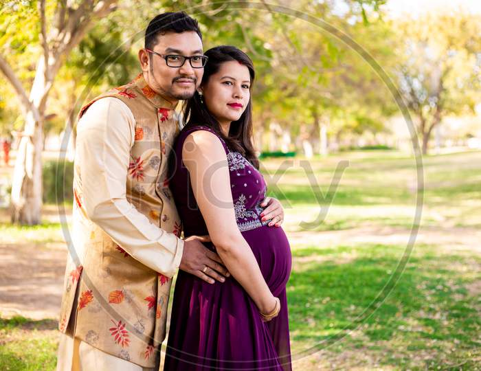 Young Asian Indian Pregnant Woman With Her Husband Wearing Traditional Outfit Standing In Park Or Garden Copy Space To Write Text.