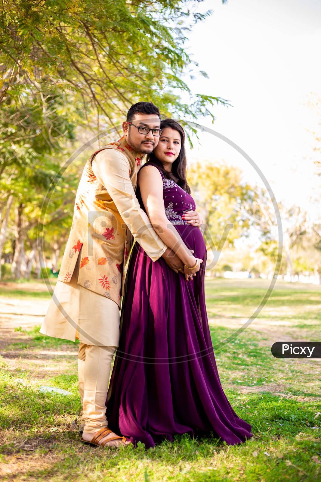 Young Asian Indian Pregnant Woman With Her Husband Wearing Traditional Outfit Standing In Park Or Garden Copy Space To Write Text.