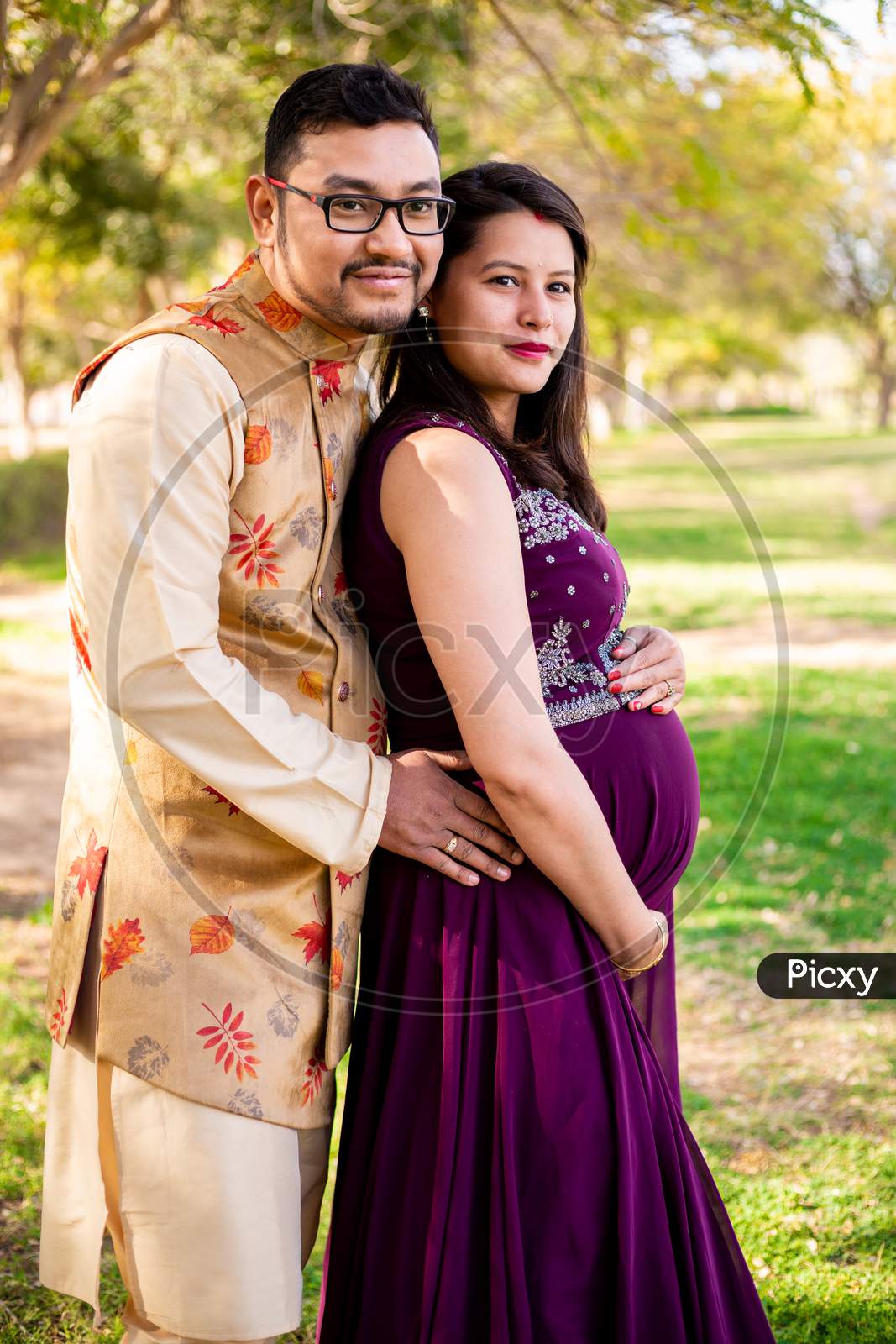 Young Asian Indian Pregnant Woman With Her Husband Wearing Traditional Outfit Standing In Park Or Garden