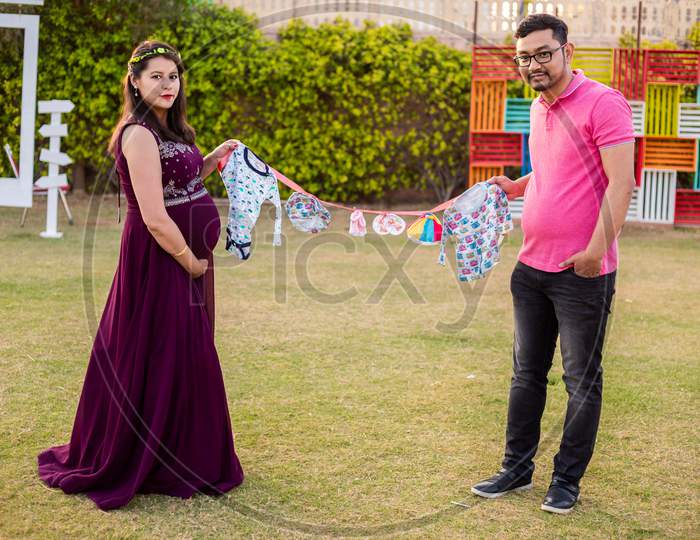 Asian Indian Pregnant Couple Holding Lots Cloth Line With Lots Of Colorful Cute Baby Outfits Standing In A Park Or Garden, Looking At Camera. Maternity Concept