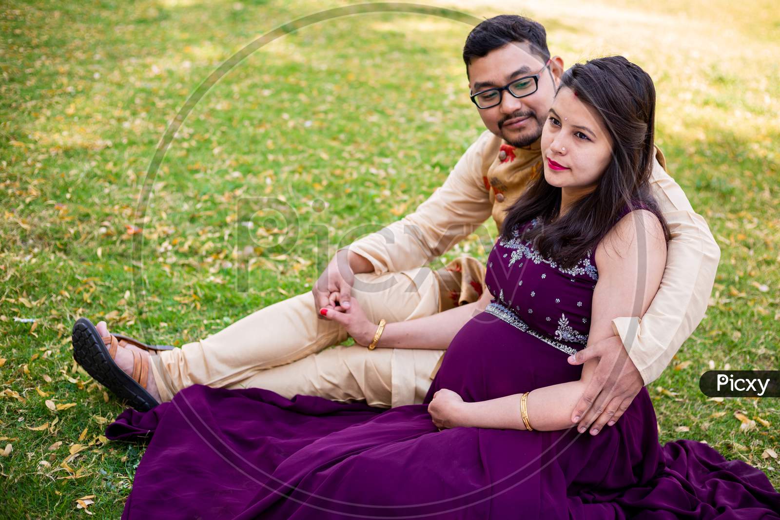 Young Asian Indian Pregnant Woman With Her Husband In Sitting Relaxing In Park Or Garden, Young Parents Looking At Camera Expecting Baby.