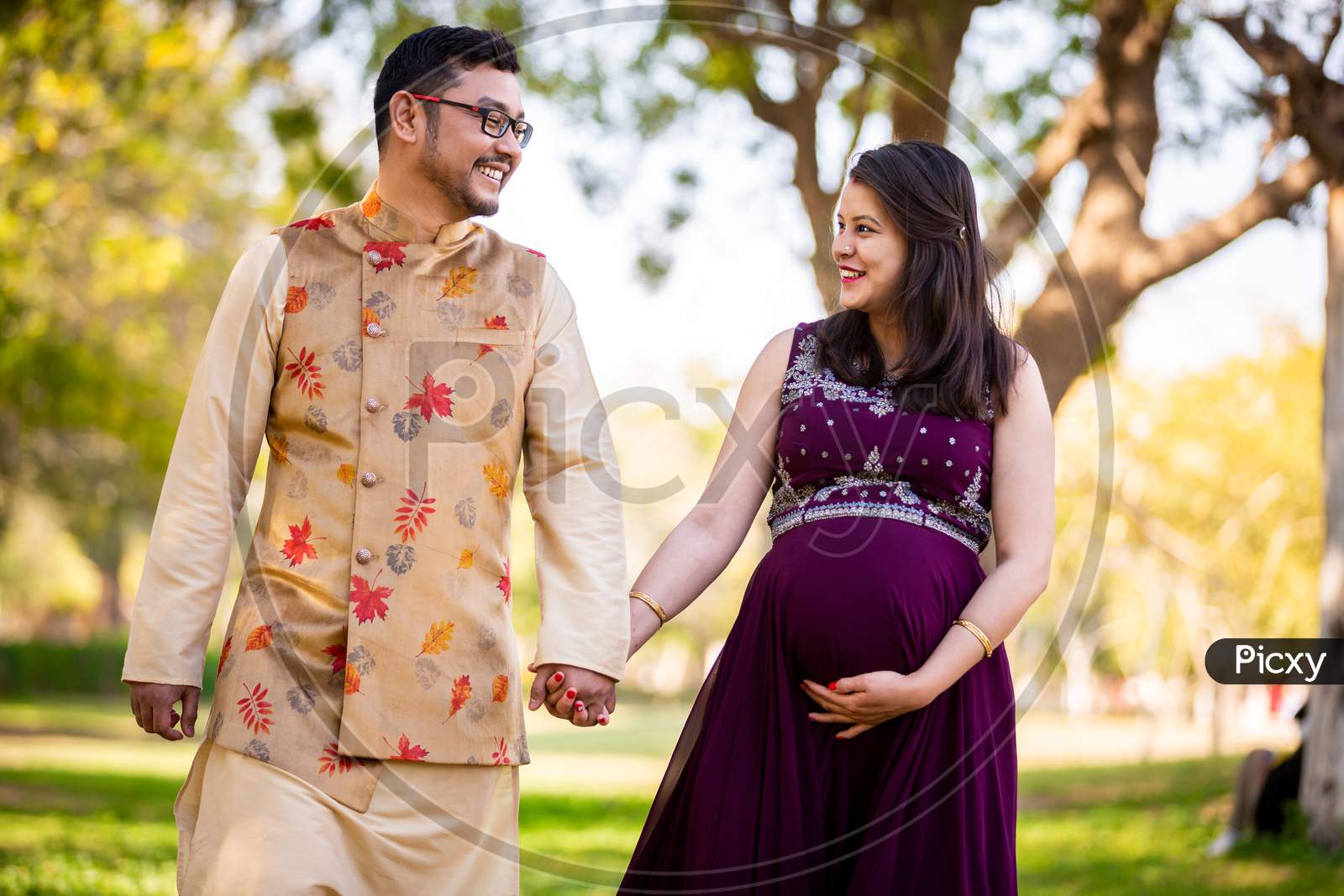 Happy Asian Indian Pregnant Woman With Her Husband Walking Outdoor In A Park Or Garden, Smiling Cheerful Young Parents Looking At Camera Expecting Baby.