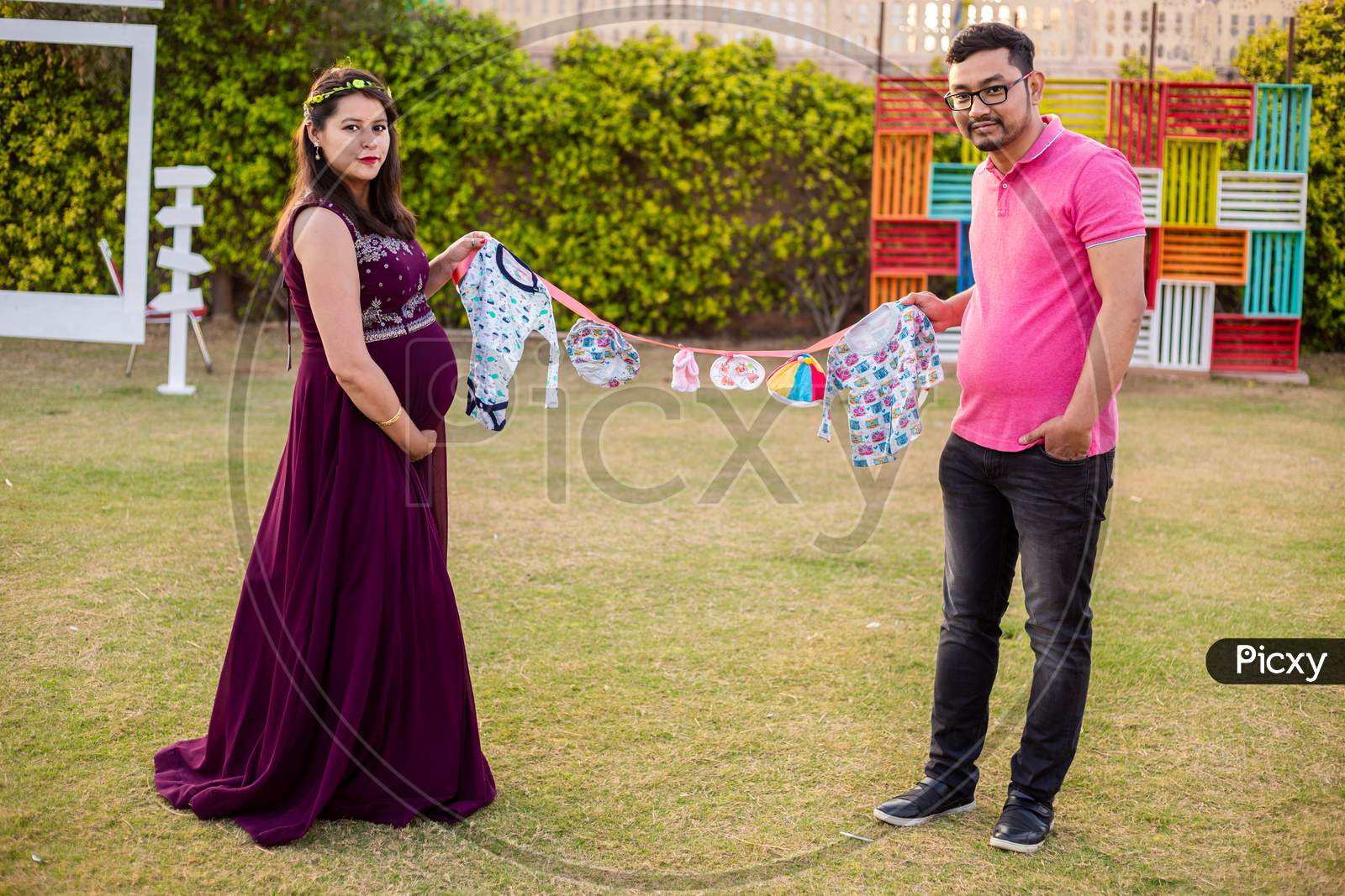 Asian Indian Pregnant Couple Holding Lots Cloth Line With Lots Of Colorful Cute Baby Outfits Standing In A Park Or Garden, Looking At Camera. Maternity Concept