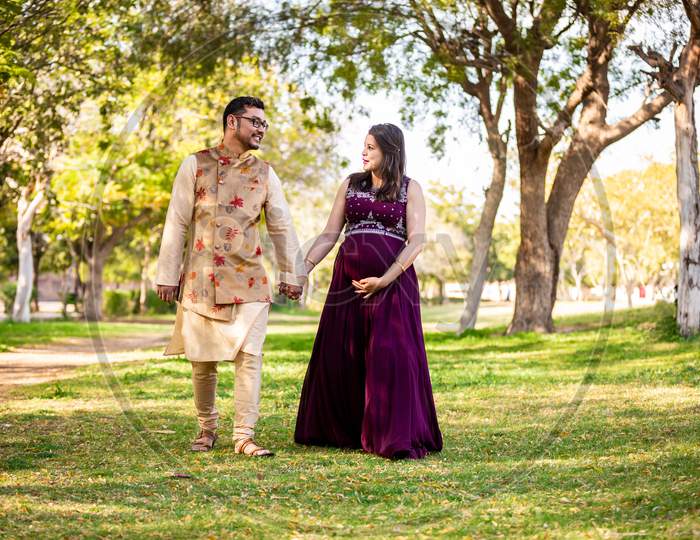 Happy Asian Indian Pregnant Woman With Her Husband In Walking Outdoor In A Park Or Garden, Smiling Cheerful Young Parents Looking At Camera Expecting Baby.