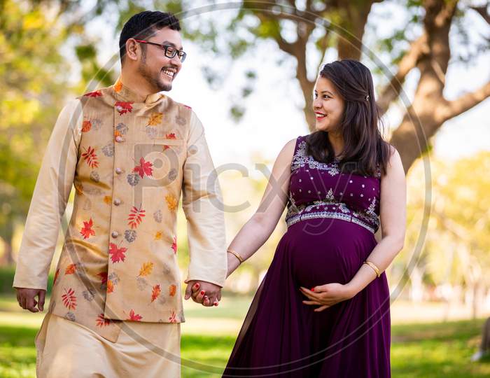 Happy Asian Indian Pregnant Woman With Her Husband Walking Outdoor In A Park Or Garden, Smiling Cheerful Young Parents Looking At Camera Expecting Baby.