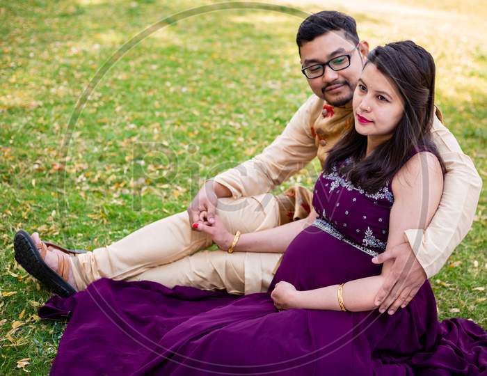 Young Asian Indian Pregnant Woman With Her Husband In Sitting Relaxing In Park Or Garden, Young Parents Looking At Camera Expecting Baby.
