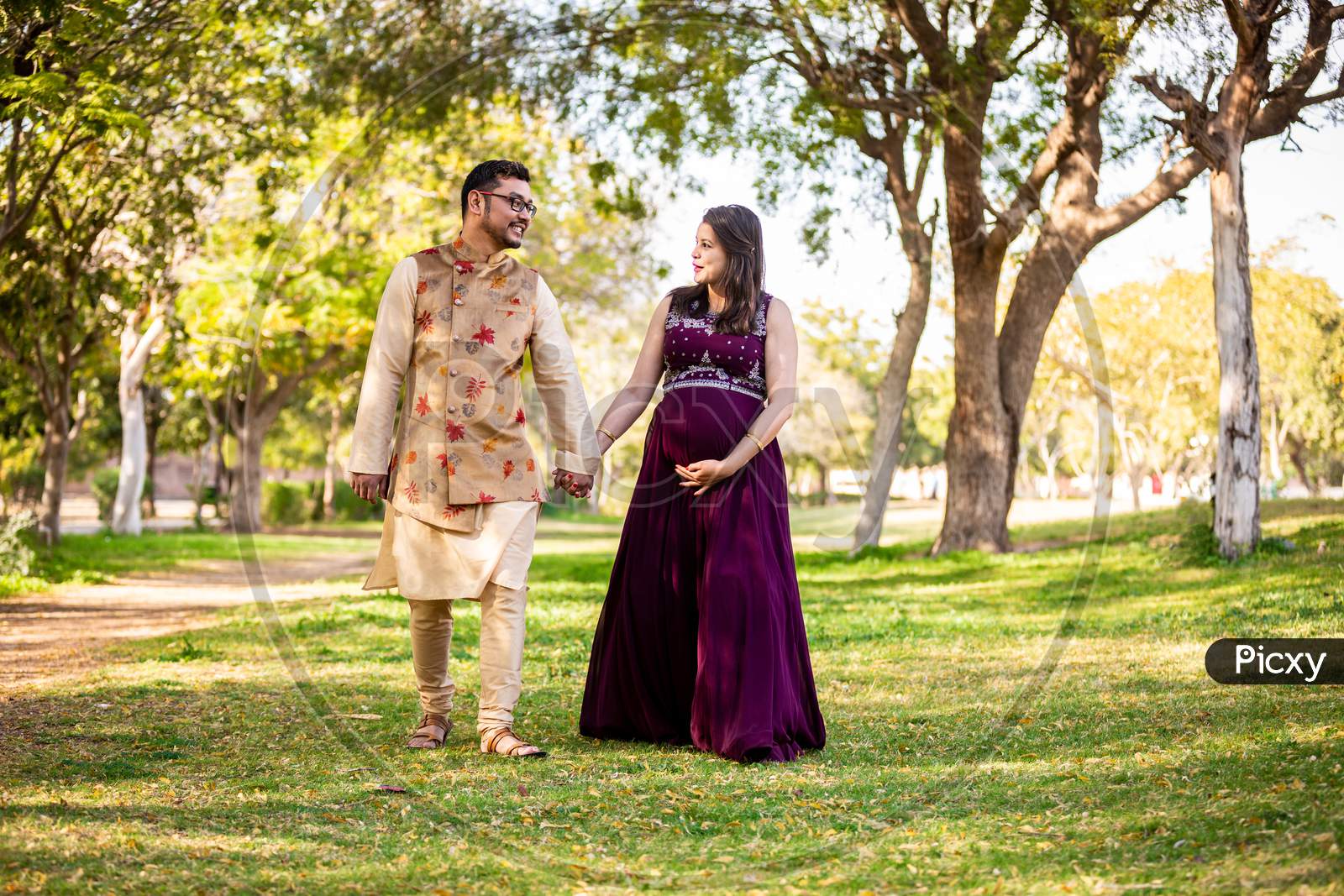 Happy Asian Indian Pregnant Woman With Her Husband In Walking Outdoor In A Park Or Garden, Smiling Cheerful Young Parents Looking At Camera Expecting Baby.