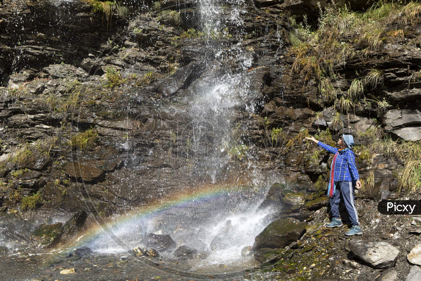 A Pretty Lady Enjoying Water Fall From A High Cliff With A Rainbow In Spiti Valley In India.