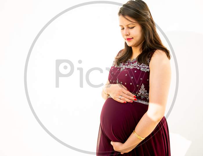 Young Asian Indian Pregnant Woman Looking At Belly Standing Against White Background Outdoor Studio Shot, Happy Female Expecting Baby, Motherhood And Pregnancy Concept, Copy Space.