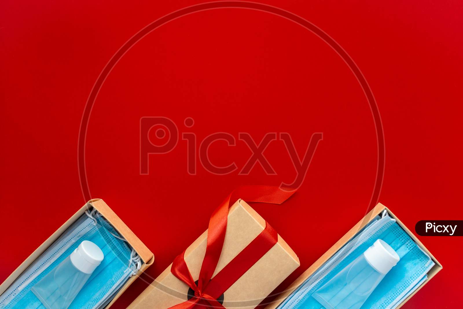 Packing A Christmas Present During Coronavirus Epidemic. Face Masks And Hydroalcoholic Gels Inside A Cardboard Box Decorated With A Bow On A Red Background.