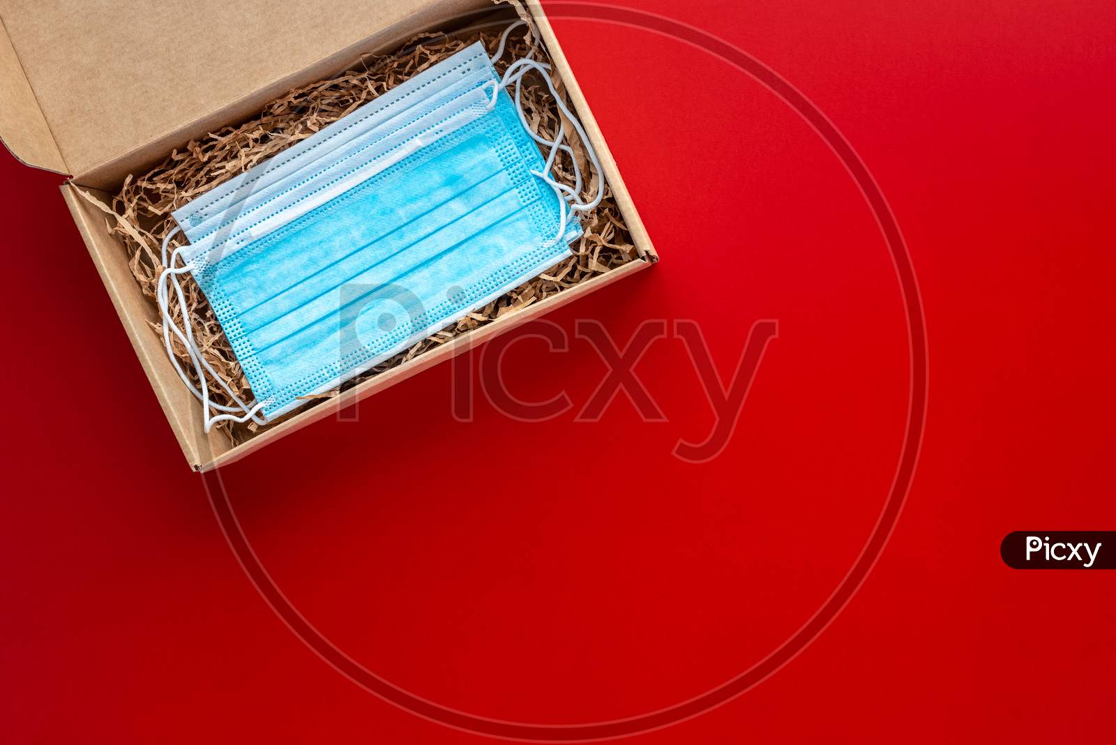 Packing A Christmas Present During Coronavirus Epidemic. Face Masks Inside A Cardboard Box On A Red Background.