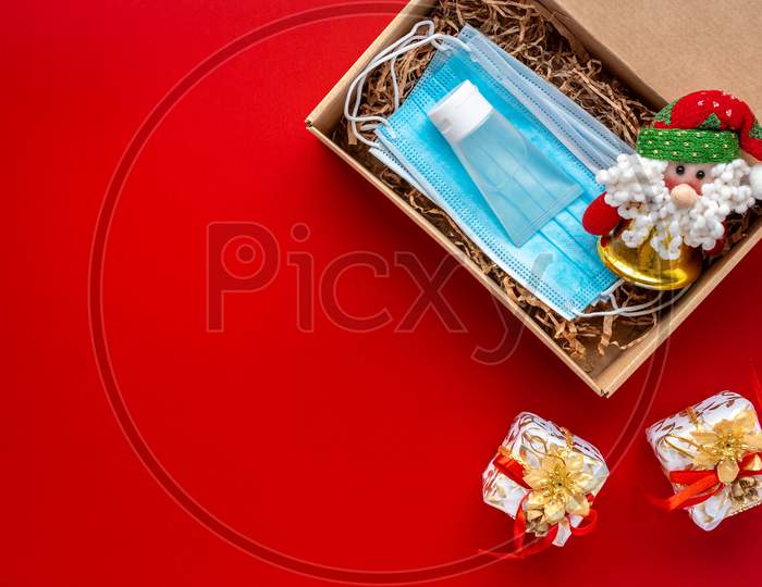 Packing A Christmas Present During Coronavirus Epidemic. Face Masks And Hydroalcoholic Gel Inside A Cardboard Box Decorated With A Bow On A Red Background.