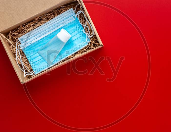 Packing A Christmas Present During Coronavirus Epidemic. Face Masks And Hydroalcoholic Gel Inside A Cardboard Box On A Red Background.