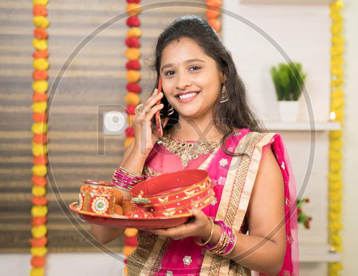 Smiling Indian Woman In Traditional Dress Holding Karva Chauth Thali Or Plate While Busy In Talking On Mobile.