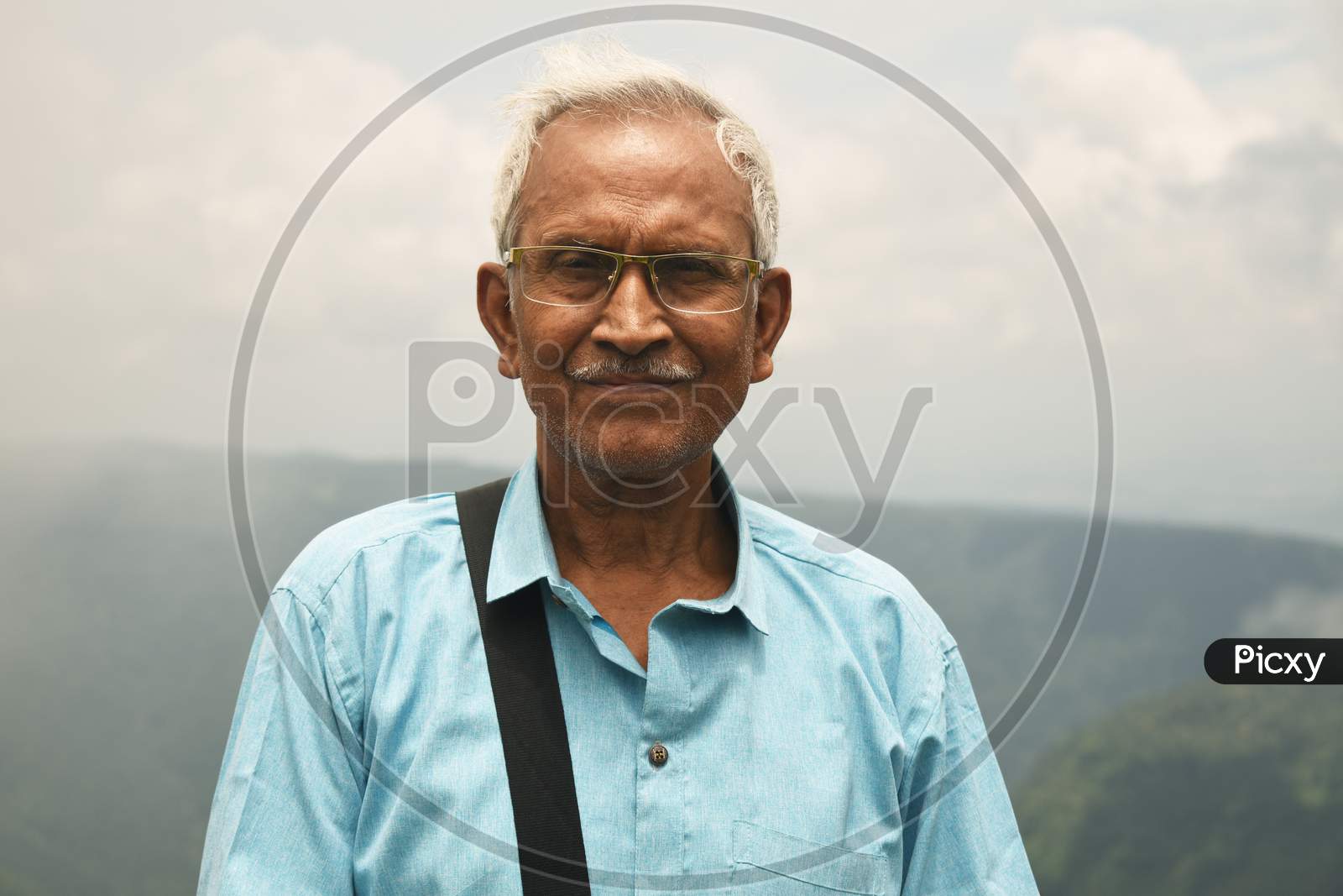 An Old Man, Wearing Spectacles And Blue Shirt, Standing Alone In Nature A Blurry Mountain Background. The Elder Person With A Side Bag On His Shoulder, Looking Towards The Camera. Copy Space For Text.