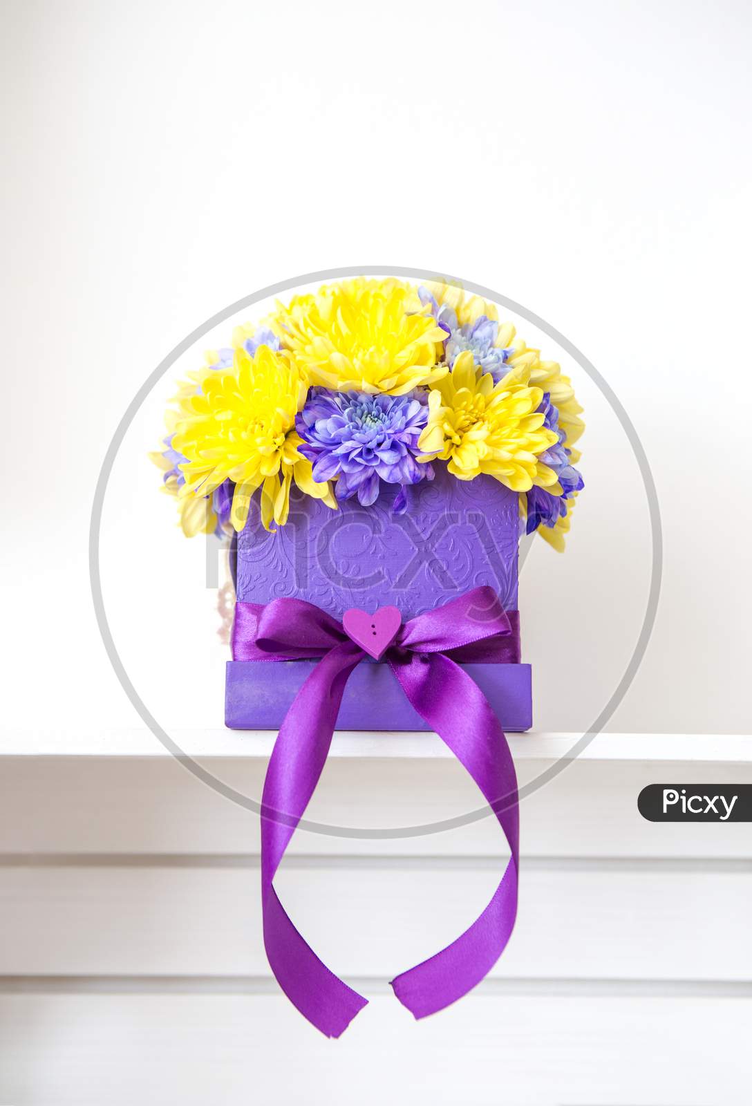Beautiful Decoration Of Chrysanthemum Flowers. The Flowers Are Yellow And Purple And Are In The Handmade Box With The Bow Also Purple. Behind Is A White Background.