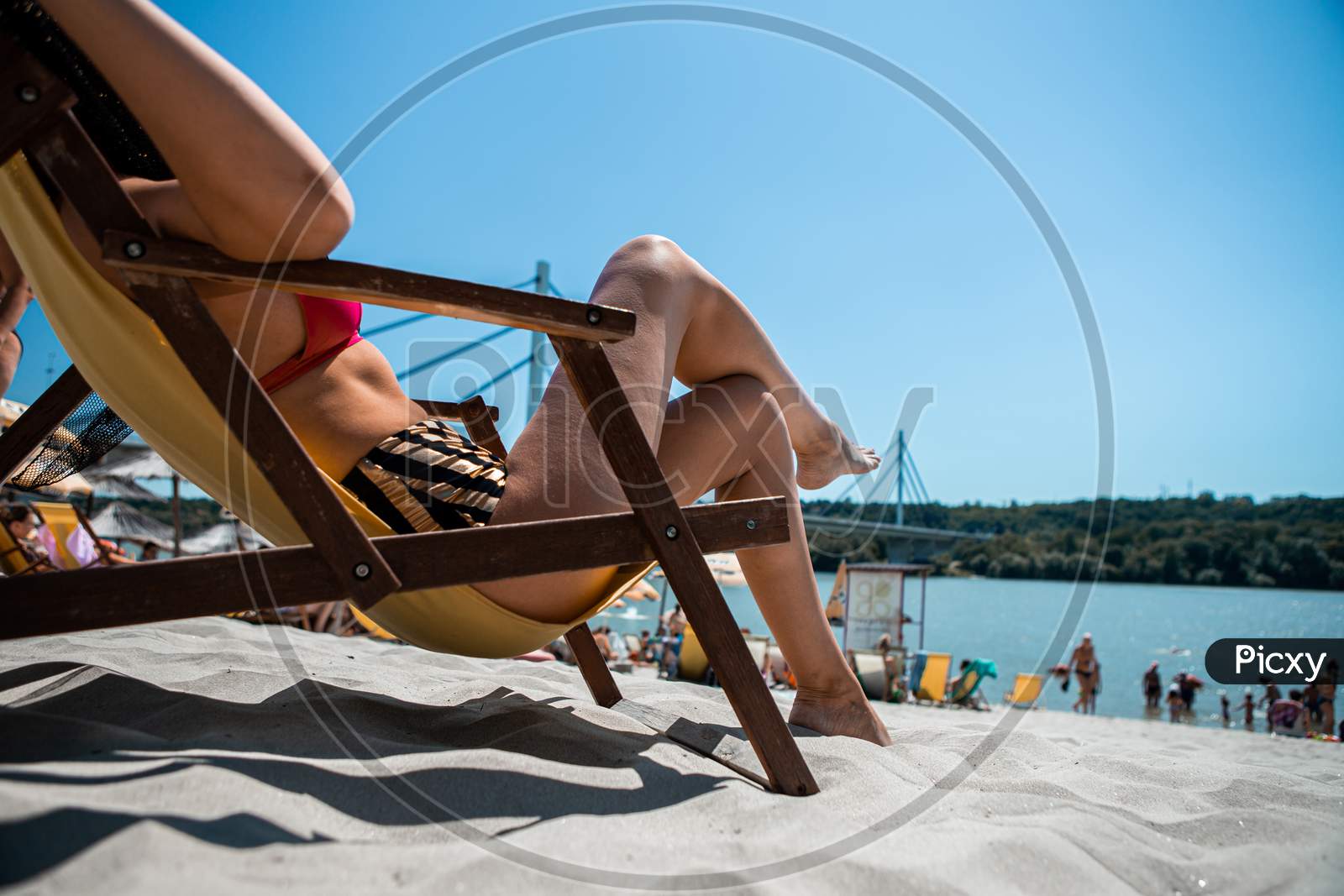 Selective Focus On The Legs Of An Attractive Young Woman Lying On A Yellow Couch On A Beach Steam. The Beach Is Sandy And Is Located On The River Bank And Behind Is A Bridge.
