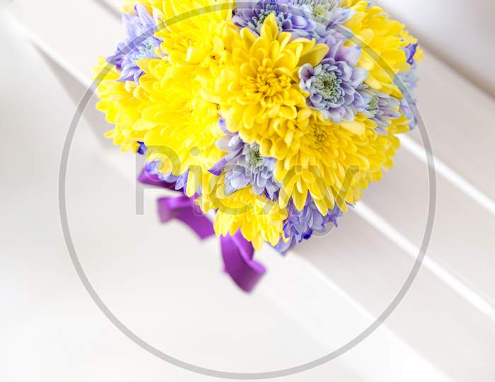 Top View Of A Decoration Of Chrysanthemum Flowers. The Flowers Are Yellow And Purple And Are In The Handmade Box With The Bow Also Purple. Behind Is A White Background.