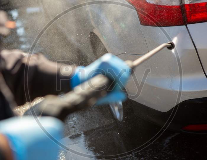 A Worker In A Car Wash Sprays A Gray Car With A Strong Jet Of Water. The Worker Wears Blue Rubber Sneakers. Car Wash. Copy Space.