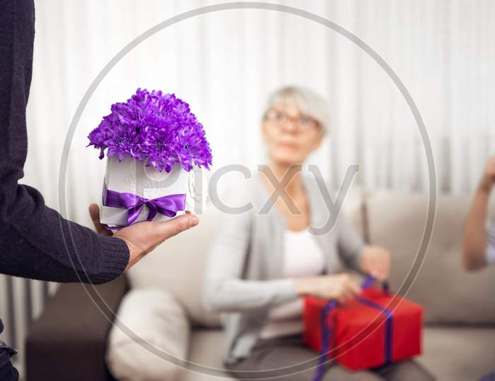 Hand Handsome Young Man And In Her Arrangement Of Purple Flowers In Front Of A Middle-Aged Blonde Lady.