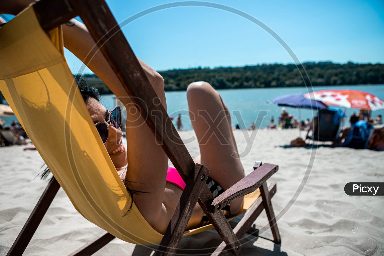 Selective Focus On The Face And Sunglasses Of An Attractive Young Woman Lying On A Yellow Deck Chair On A Beach Steam. The Beach Is Sandy And Located On The River Bank.