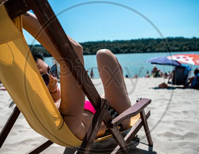 Selective Focus On The Face And Sunglasses Of An Attractive Young Woman Lying On A Yellow Deck Chair On A Beach Steam. The Beach Is Sandy And Located On The River Bank.