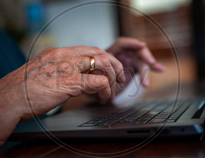 Focus On The Hand Of An Elderly Senior Lady Using A Laptop. She Searches For Something On The Internet. On His Hand Is An Old Engagement Gold Ring.