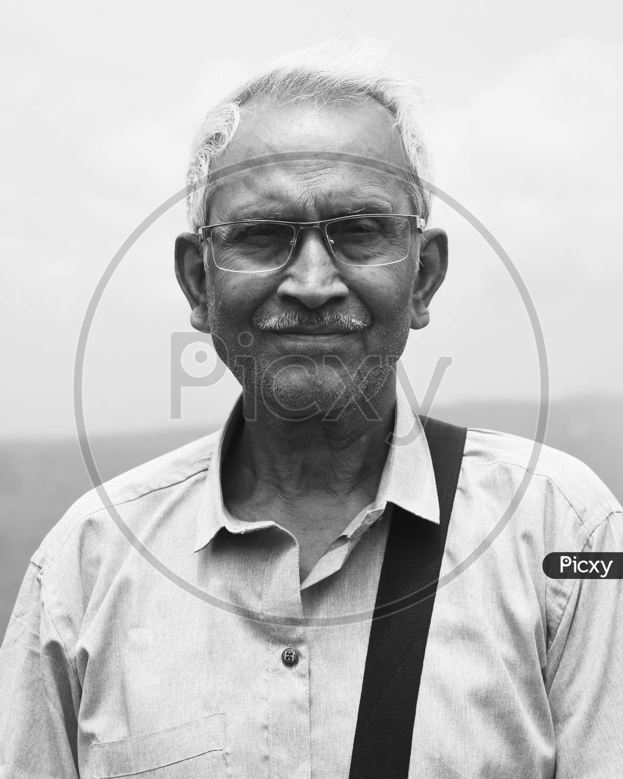 Monochrome Portrait Of An Old Man, Wearing Spectacles And Blue Shirt, Standing Alone In Nature A Blurry Mountain Background. The Elder Person With Side Bag On His Shoulder, Looking Towards The Camera.