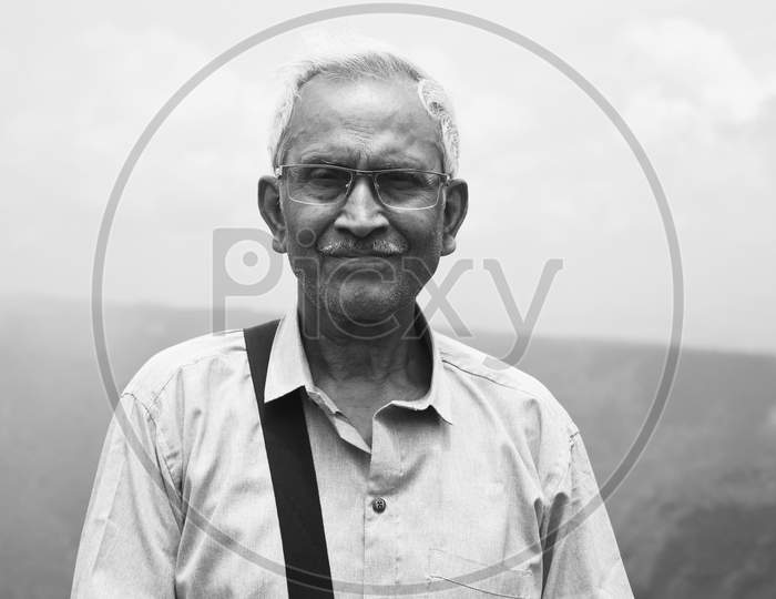 Monochrome Portrait Of An Old Man, Wearing Spectacles And Blue Shirt, Standing Alone In Nature A Blurry Mountain Background. The Elder Person With Side Bag On His Shoulder, Looking Towards The Camera.
