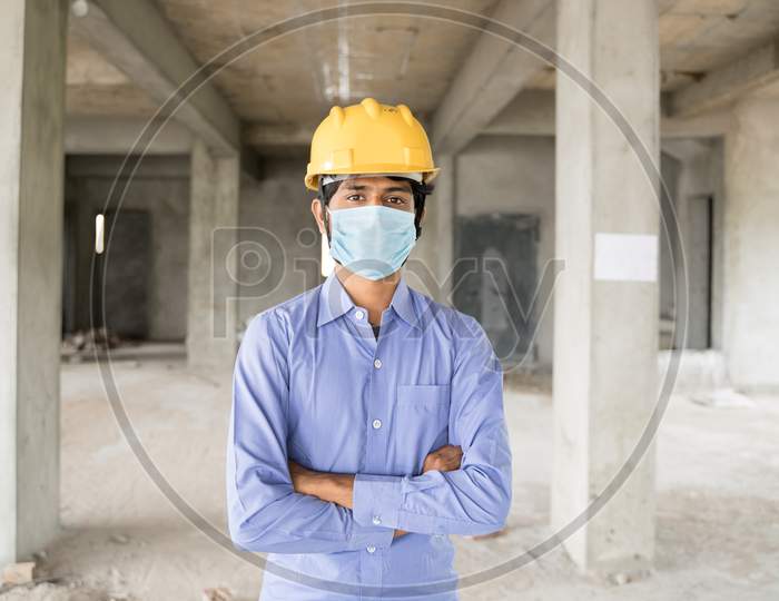 Concept Of Back To Work, Opening Of Construction Sites After Covid-19 Pandemic - Portrait Of Confident Construction Worker In A Construction Helmet Arms Crossed With Medical Mask At Site.