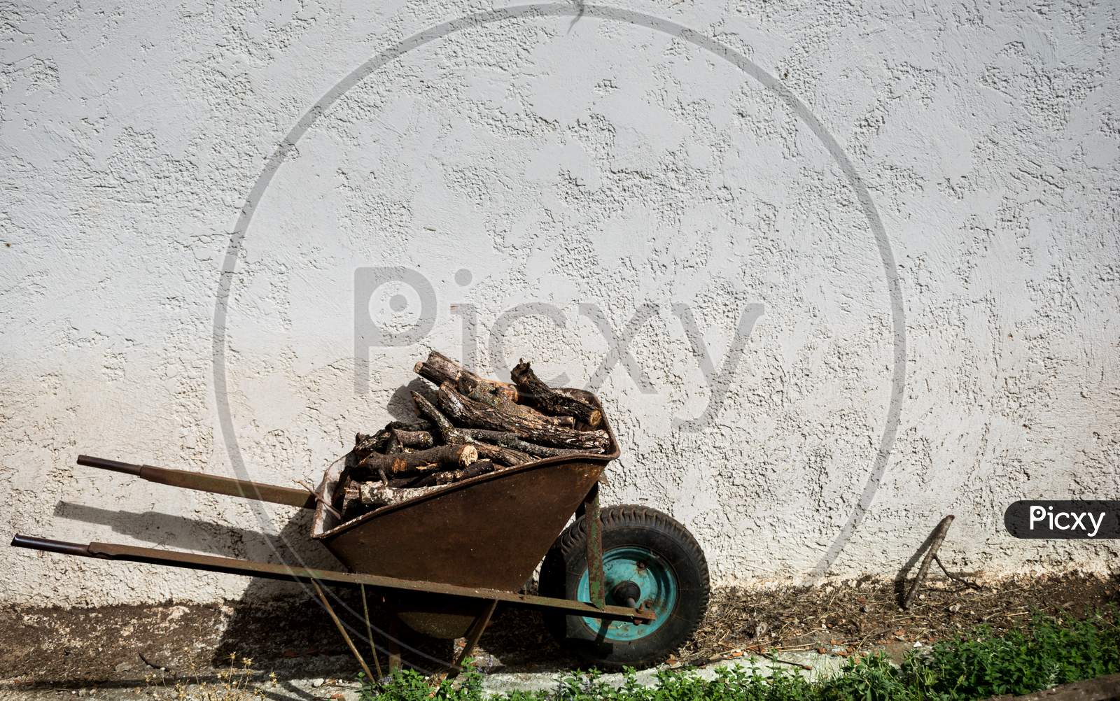 A Rusty Wheelbarrow Loaded With Firewood Stands In Front Of A White Wall Background.