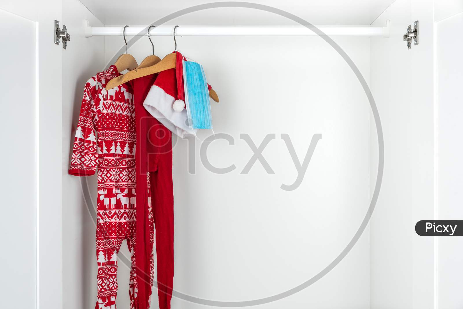 White And Red Christmas Pajamas, Hat, Tights And Face Mask Hanging On Wooden Hangers Inside A White Closet. Holidays During Coronavirus Pandemic.