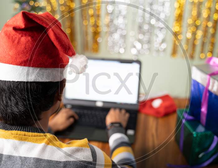 Shoulder Shot Of Young Man Busy Working On Laptop With Santa Hat During Christmas Or New Year Eve - Concept Of Work From Home During Holiday Season Due To Coronavirus Or Covid-19 Pandemic