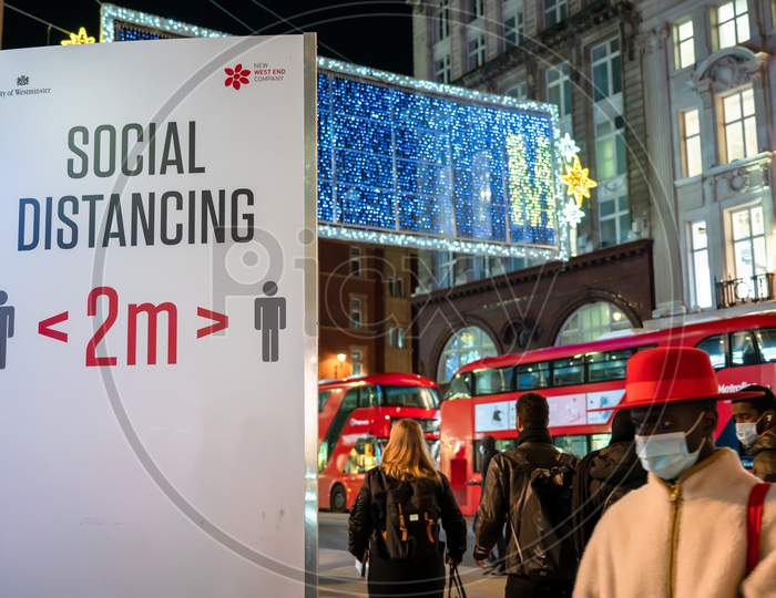 Covid 19 Social Distancing Sign And Oxford Street Illuminated Christmas Decorations With A Black Man In Face Mask And Red Hat