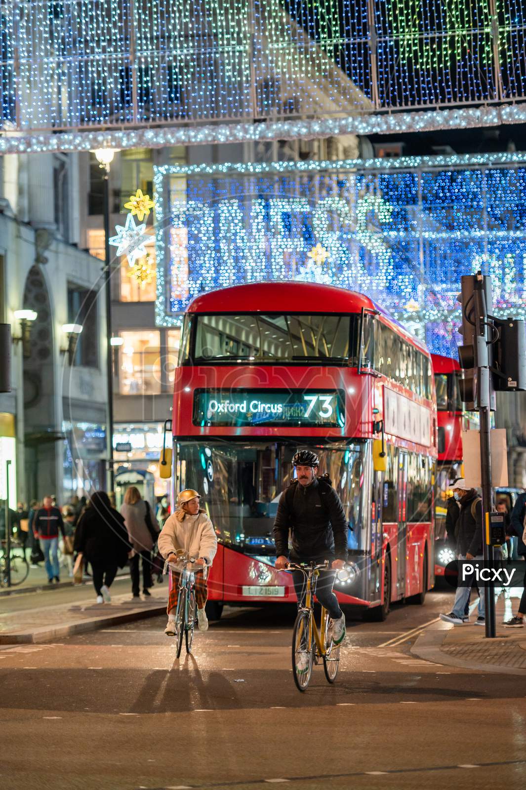 Cyclists Ahead Of A Red London Double Decker Bus With Oxford Street Illuminated Christmas Decorations In The Background