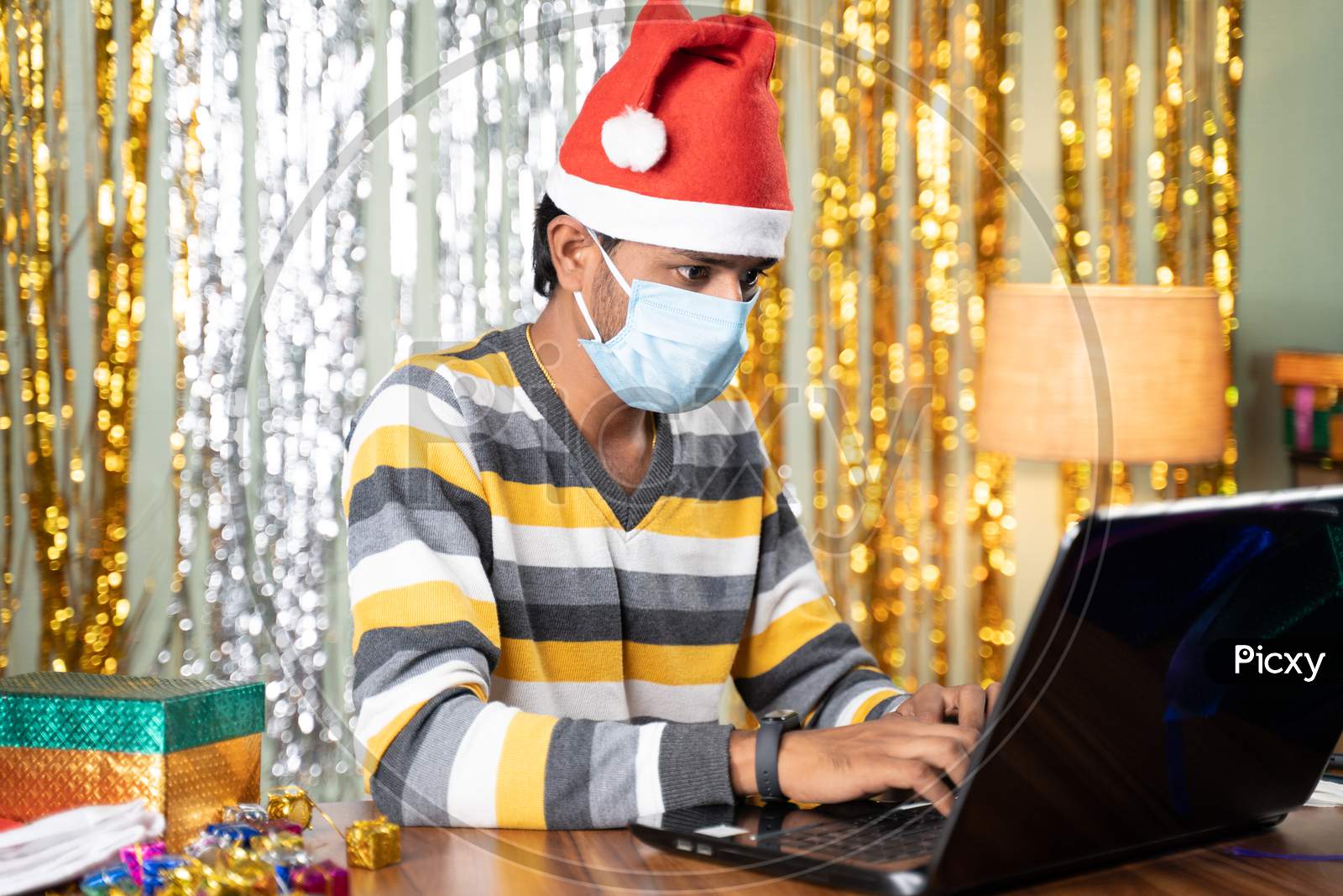 Young Man In Medical Mask Busy Working On Laptop During Christmas Or New Year Celebration Eve With Decorated Background And Gifts In Front - Concept Of Work From Home During Holydays.