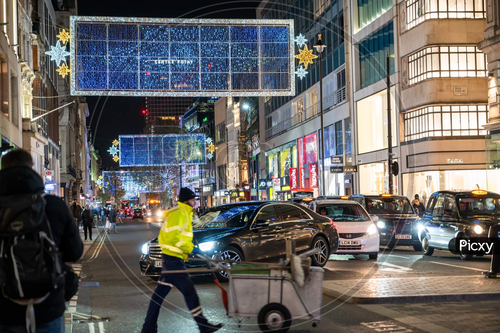 A Street Cleaner Pushes A Cart Between Traffic Beneath The Oxford Street Christmas Decorations At Night