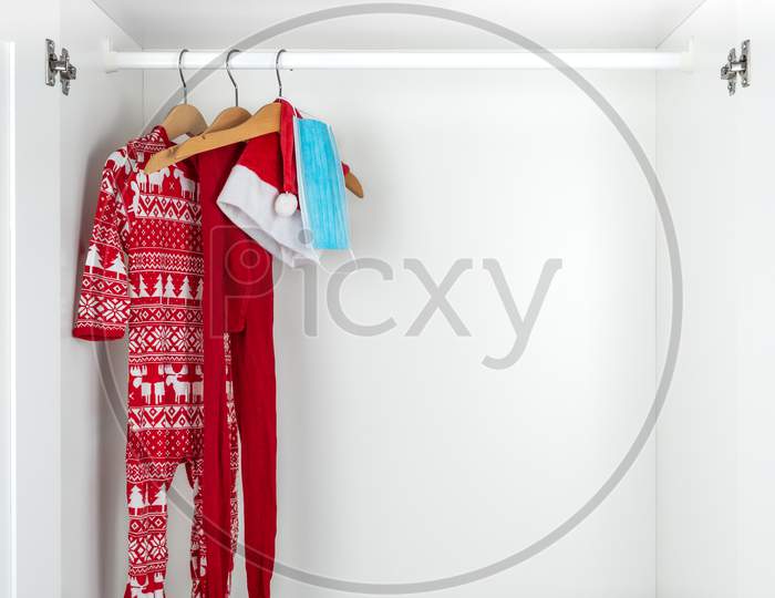 White And Red Christmas Pajamas, Hat, Tights And Face Mask Hanging On Wooden Hangers Inside A White Closet. Holidays During Coronavirus Pandemic.