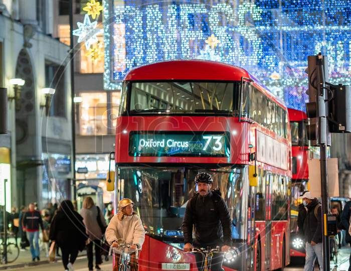 Cyclists Ahead Of A Red London Double Decker Bus With Oxford Street Illuminated Christmas Decorations In The Background