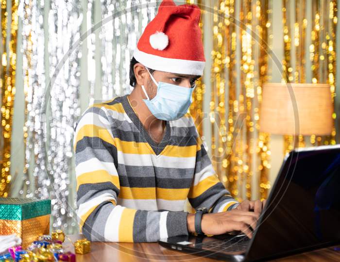 Young Man In Medical Mask Busy Working On Laptop During Christmas Or New Year Celebration Eve With Decorated Background And Gifts In Front - Concept Of Work From Home During Holydays.
