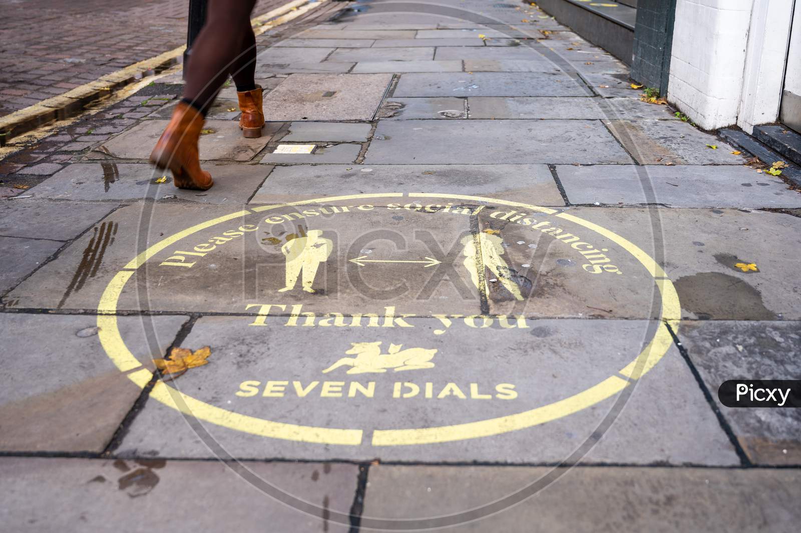 A Woman'S Legs Walk Past A Yellow Covid 19 Social Distancing Sign Painted On The Pavement In Covent Garden