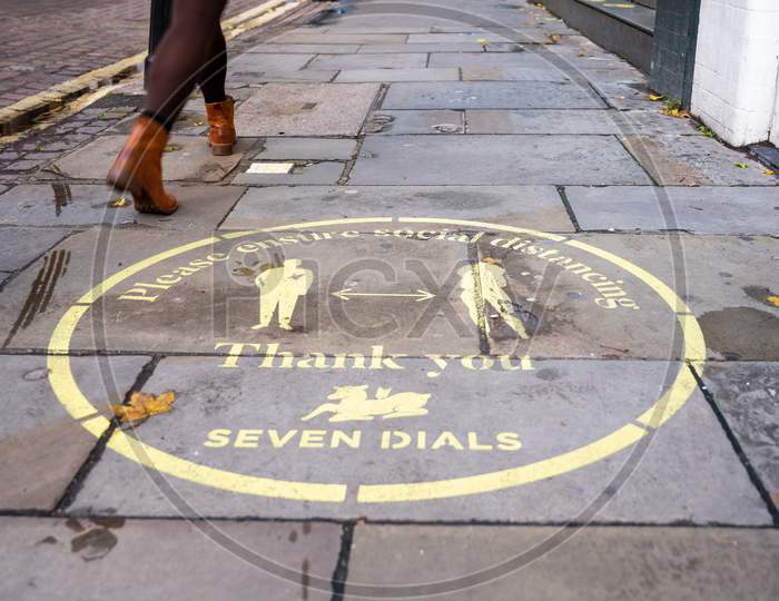 A Woman'S Legs Walk Past A Yellow Covid 19 Social Distancing Sign Painted On The Pavement In Covent Garden