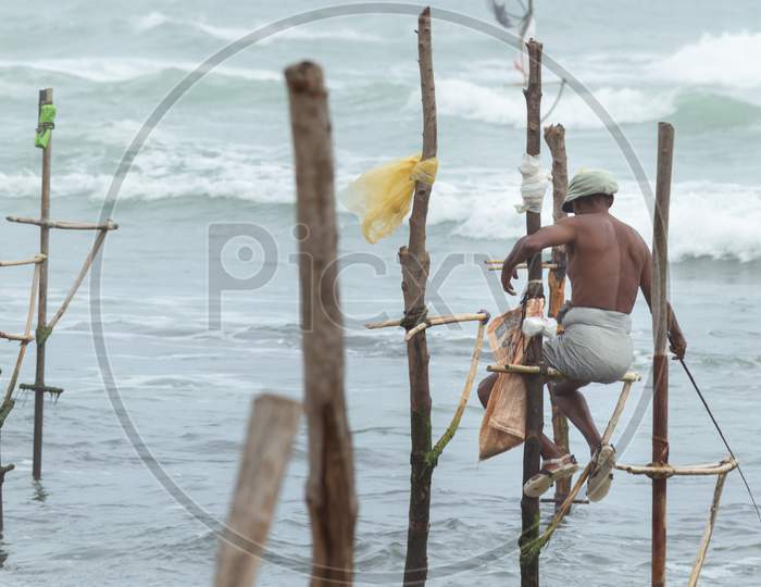Old Stilt Fisherman With His Wooden Rod Facing Back To Camera Angel, Fishing In A Traditional Unique Method In Sri Lankan Culture, Sunny Bright Evening On The Beach.