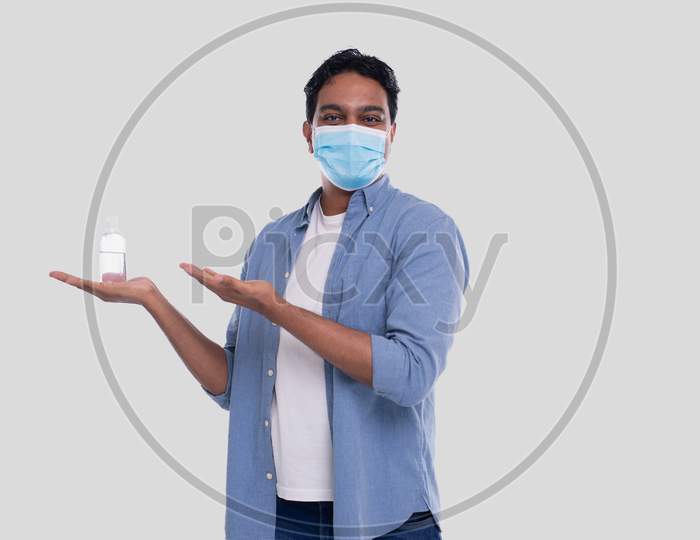 Man Showing Antiseptic On Hand And Pointing At It Wearing Medical Mask Isolated. Indian Man Wearing Medical Mask